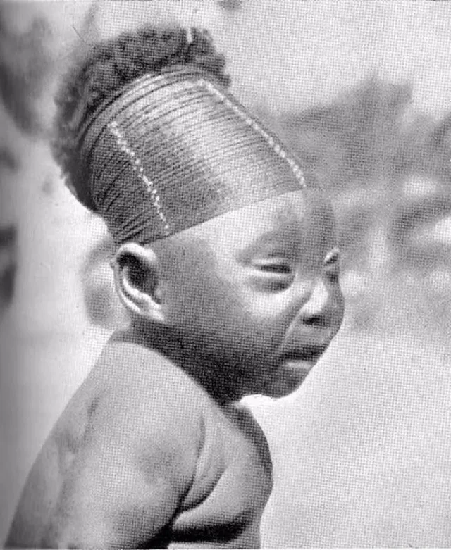 A-young-Mangbetu-girl-holding-her-baby-sibling-whose-head-wrapping-reflects-the-ongoing-pract...webp